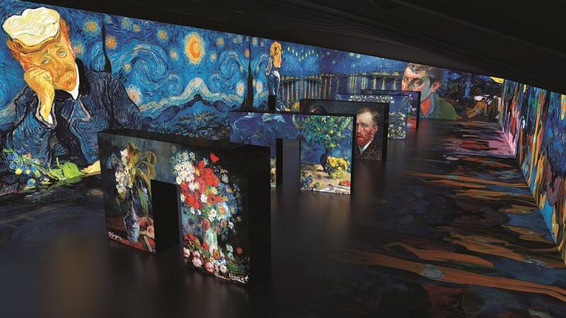 The image shows an immersive art exhibit featuring the works of Vincent van Gogh, with large-scale projections of his paintings on the walls and freestanding displays.