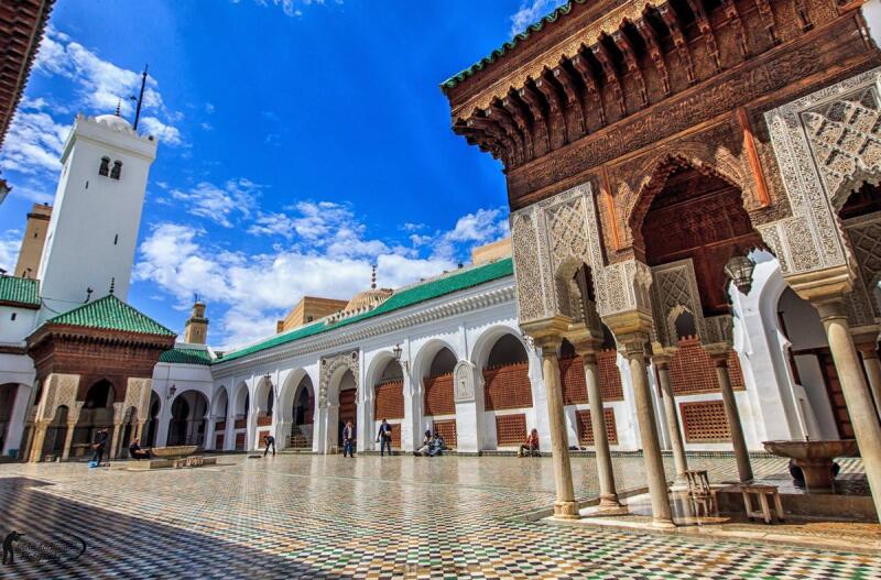 The image shows the courtyard of Al-Qarawiyyin Mosque in Fez, Morocco, featuring a tiled floor, white arched corridors, and a tall minaret under a blue sky with scattered clouds.