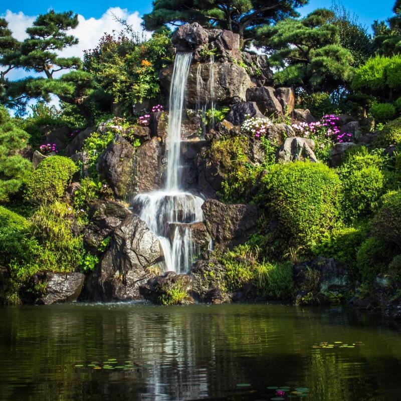 A waterfall cascades over rocks surrounded by greenery into a pond.