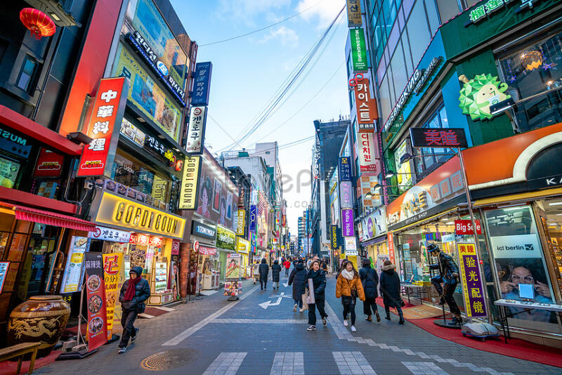 A street view in a bustling urban area, lined with various shops and restaurants adorned with colorful signs and advertisements, with pedestrians walking along the sidewalk.