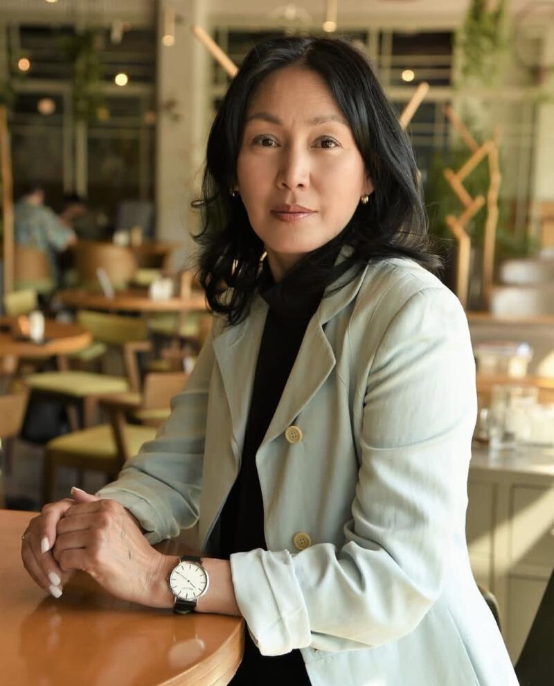 A woman with dark hair is sitting at a table, wearing a light green jacket over a black turtleneck, and a watch on her left wrist.