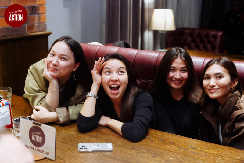 Four smiling young women are sitting at a wooden table, with a phone and a menu with the text "ACTION" on it, in a room with a brick wall and a burgundy leather sofa.