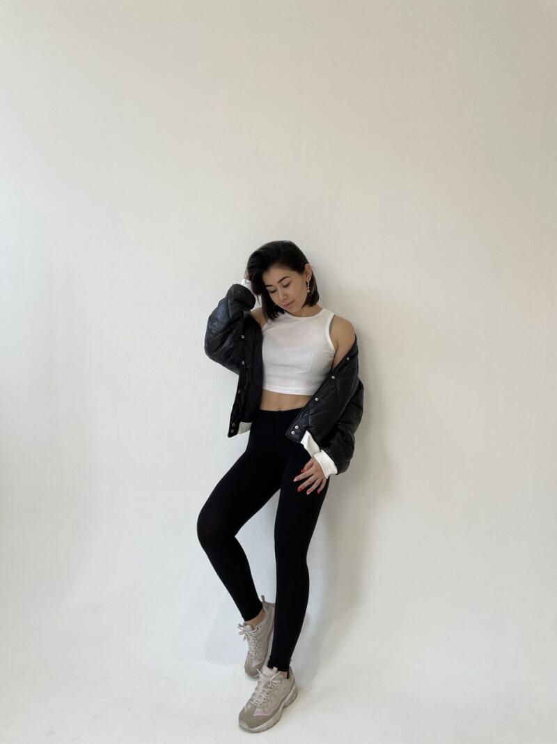 A woman with short dark hair is wearing a white top, black leggings, a black jacket, and gray sneakers while posing against a white wall.