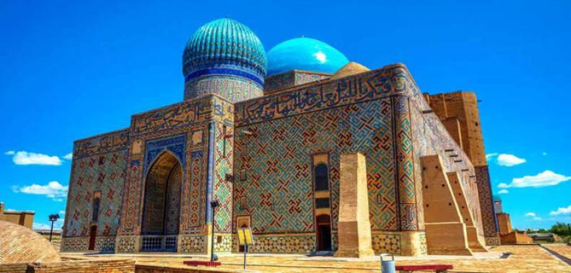 The image shows the exterior of a building with two large, rounded domes, one of which is decorated with a mosaic pattern. The building is intricately patterned with geometric shapes and Arabic script and has a large arched entrance.
