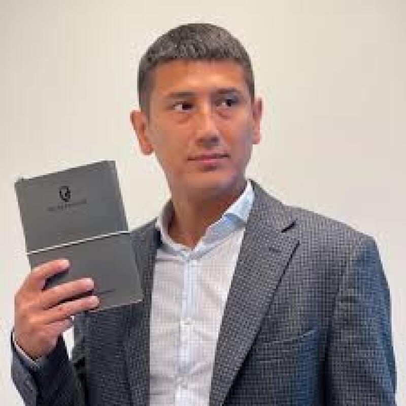 A man in a grey blazer and light blue shirt is holding up a dark grey book with a logo and text on it.