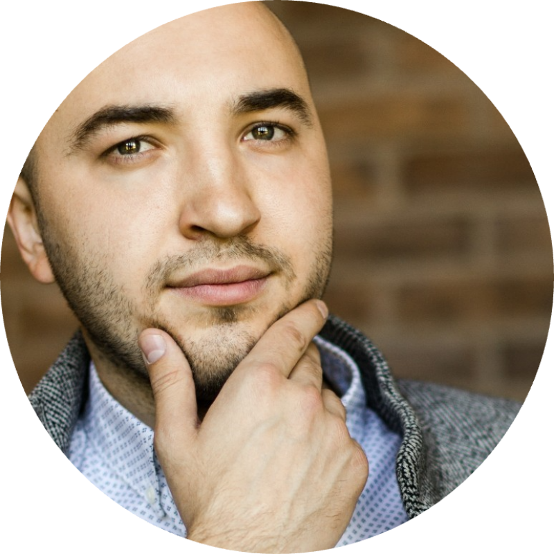 A man with a light beard and green eyes is holding his chin, wearing a blue patterned shirt and a gray jacket, in front of a brick wall.