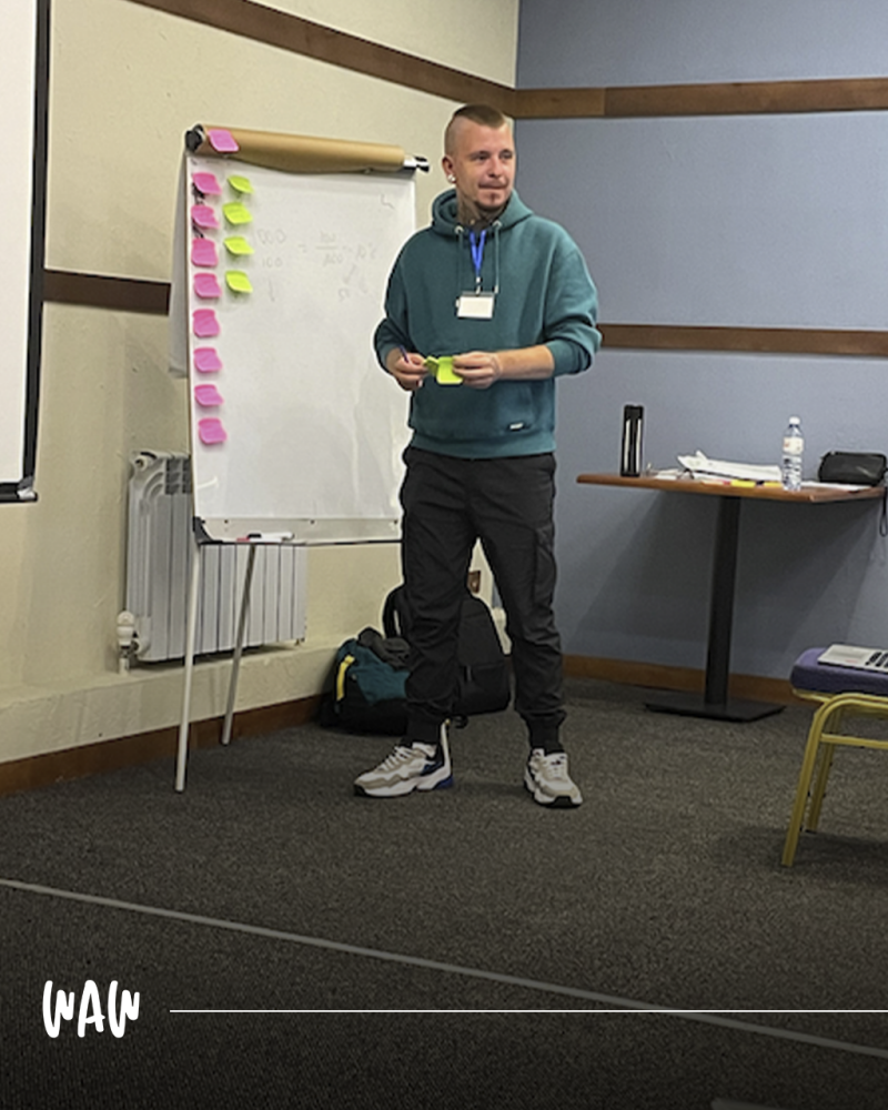 Мужчина в толстовке стоит возле белой доски, покрытой стикерами, и держит желтый стикер; на заднем плане стол с бутылкой воды и другими предметами.