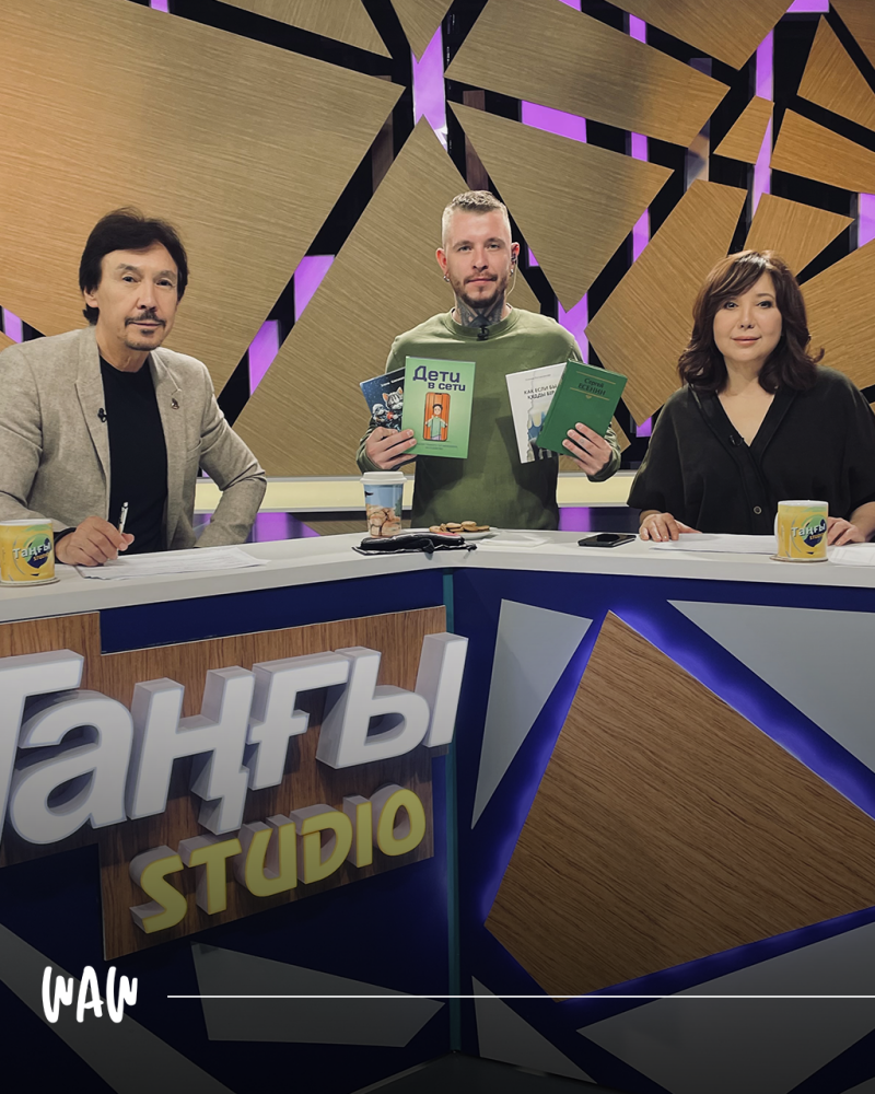 Три человека сидят за столом с логотипом "Tanby Studio", держа в руках книги и бумаги.