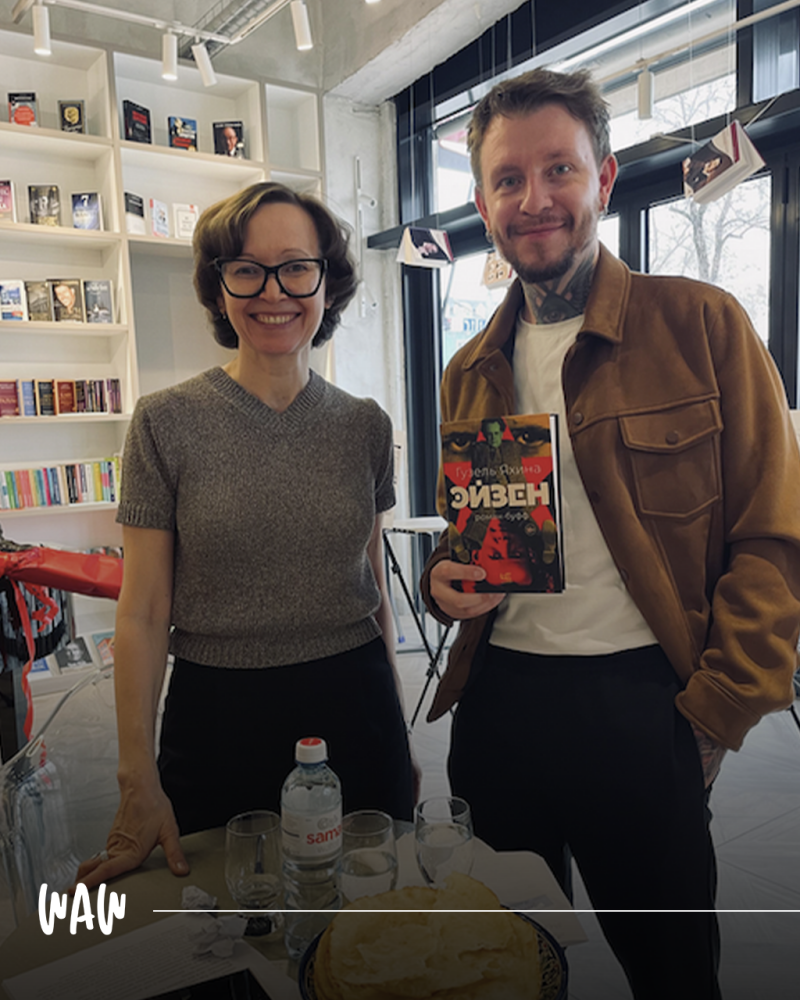 Два человека позируют вместе, один держит книгу под названием «Дизель» на русском языке, в комнате с книжными полками.