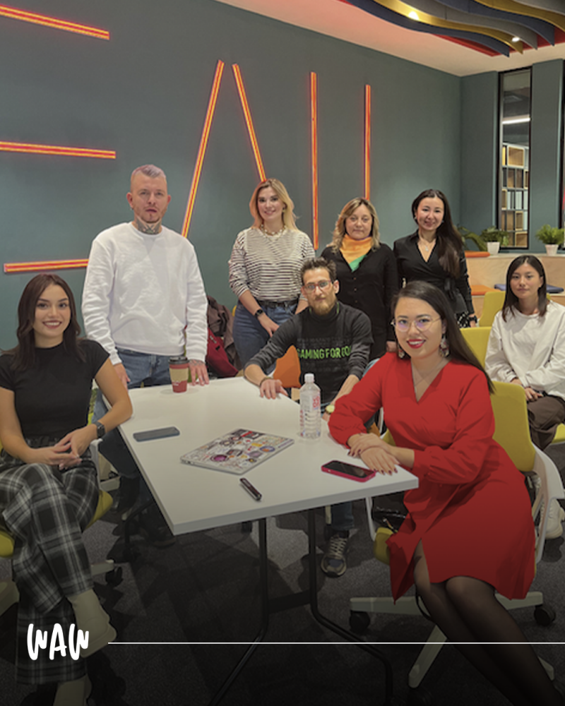 Группа из восьми человек позирует за белым столом в современном офисе с неоновыми настенными светильниками, представляя разнообразную группу людей в разной одежде, включая женщину в красном платье и мужчину с татуировками на шее.