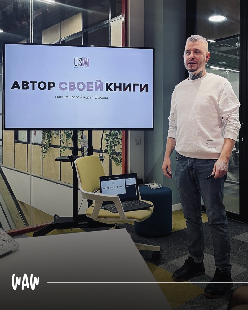 Мужчина в белом свитшоте и джинсах стоит рядом с экраном, на котором отображается текст "АВТОР СВОЕЙ КНИГИ" и "мастер-класс Андрея Орлова", а на стоящем рядом стуле лежит ноутбук, на котором также виден этот текст.