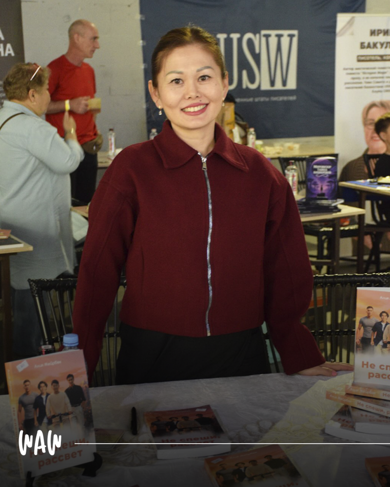 Женщина в красной куртке стоит за столом с книгами, улыбаясь в камеру; на заднем плане видны люди и баннер с текстом 'USW'.