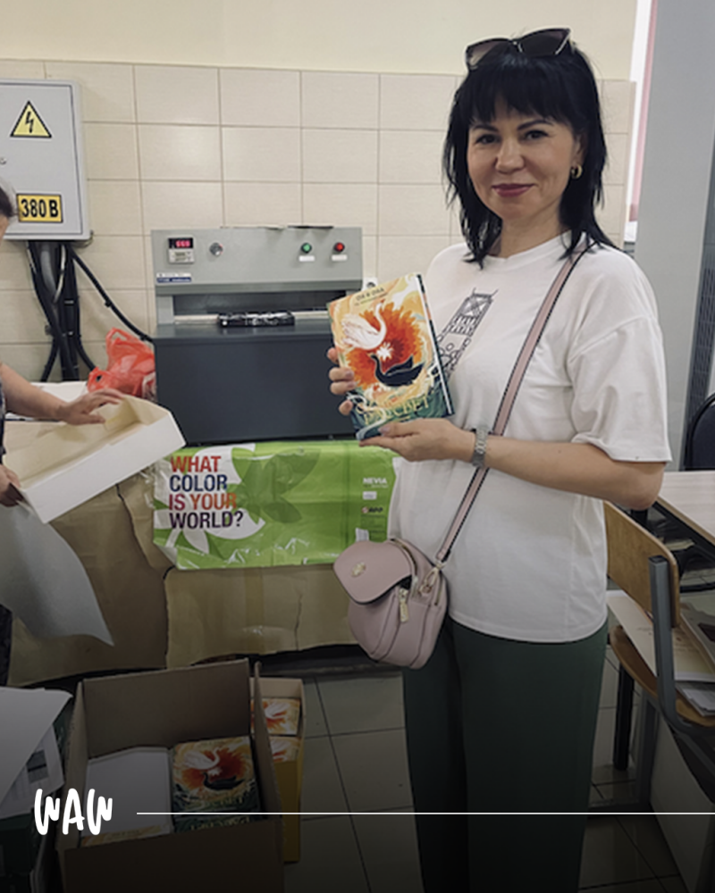 Женщина с солнечными очками на голове держит книгу с изображением лебедя, стоя перед столом с коробками и вывеской с надписью "WHAT COLOR IS YOUR WORLD?"