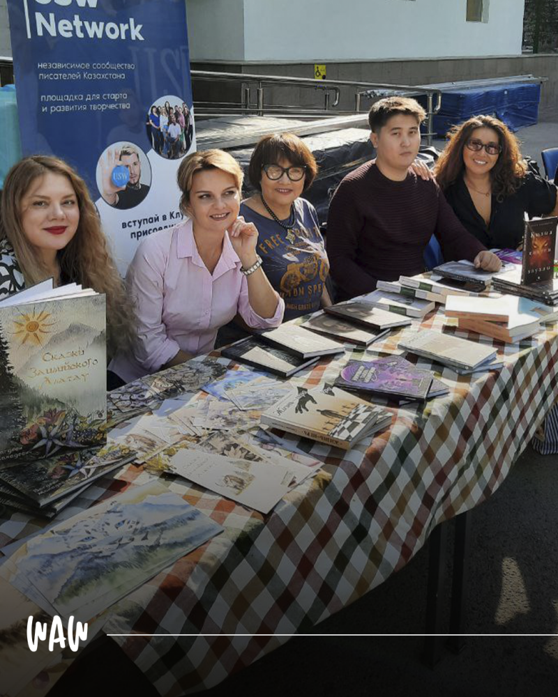 Четыре человека сидят за столом, покрытым клетчатой скатертью, на котором разложены книги и картины для продажи; на заднем плане баннер с надписью 'CSW Network, независимое сообщество писателей Казахстана, площадка для старта и развития творчества', а также приглашение вступить в клуб.