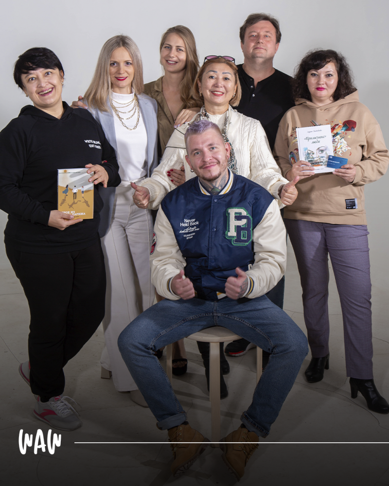 Группа из семи человек позирует для фотографии, один мужчина сидит на табурете впереди. Несколько человек держат книги, в том числе книгу под названием «ПРИЛИЧНОГО ЛЮДИ».