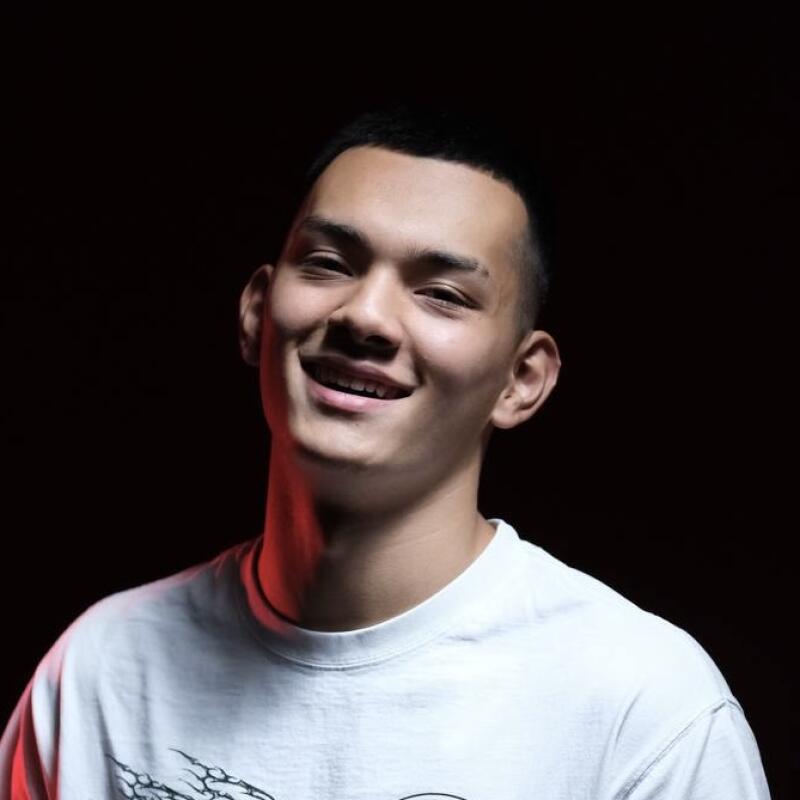 A smiling young man with short dark hair and a white t-shirt stands against a dark background.