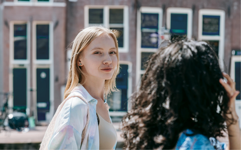 A blonde woman looks at the camera while standing outside with another woman with curly hair.