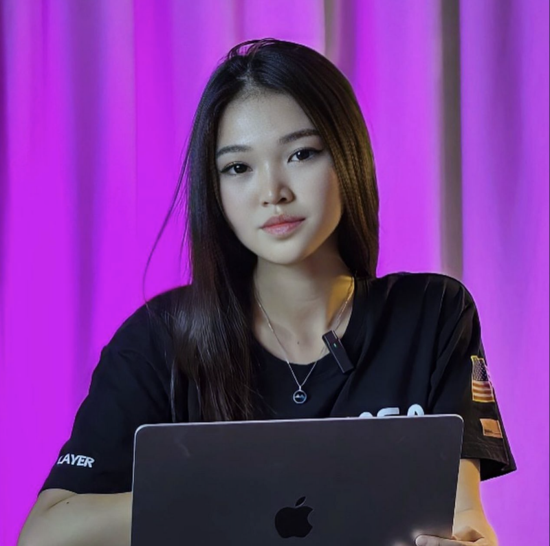 A woman with long dark hair is holding a laptop in front of her, wearing a black T-shirt with a small American flag patch on the sleeve, against a purple backdrop.