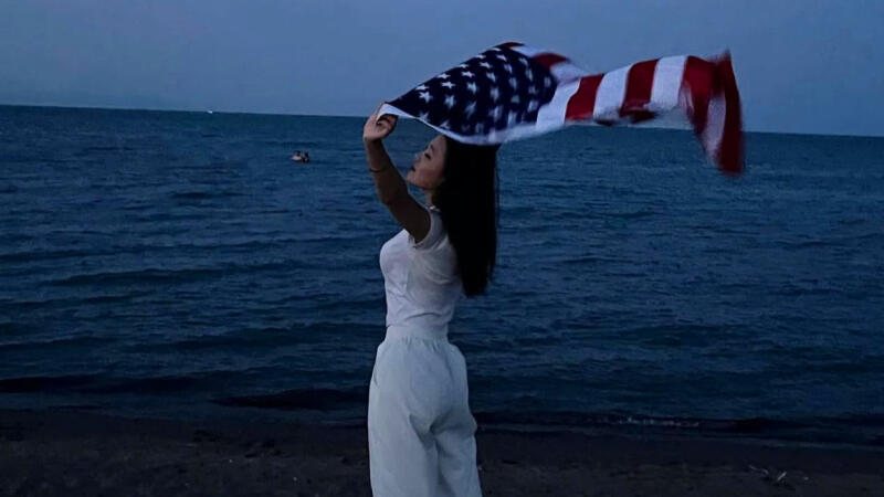 A person in white clothing stands on a beach holding up an American flag against a backdrop of the sea and a dusky sky.