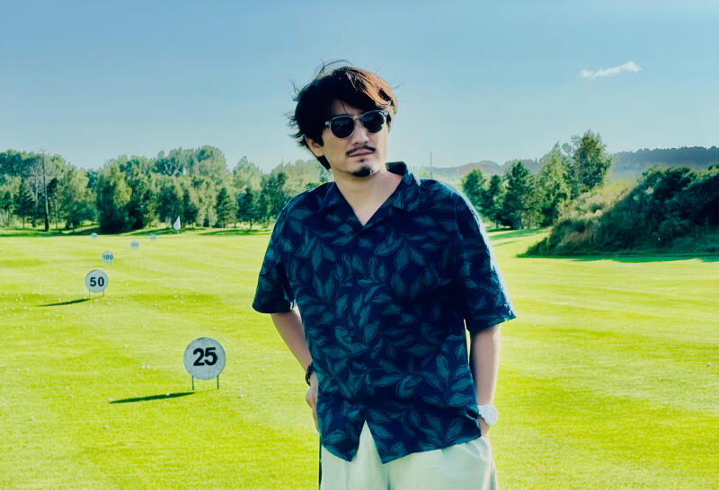 A man in sunglasses and a blue leafy shirt stands on a golf course, with markers labeled '25,' '50,' and '100' visible in the background.