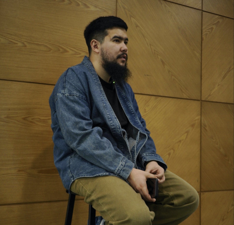 A man with a beard is sitting on a stool, wearing a blue denim jacket, a black t-shirt, and khaki pants, holding a phone in his hands, against a wood-patterned wall.