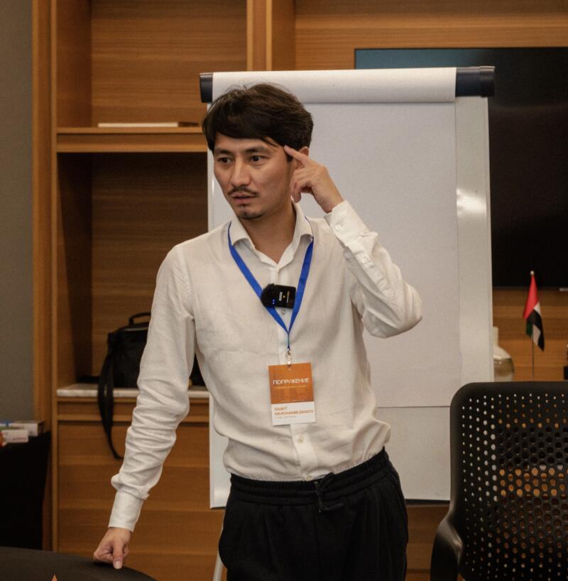 A man with a mustache, wearing a white shirt and black pants, is gesturing with his hand to his head in front of a whiteboard; he is wearing a badge that reads "ПОГРУЖЕНИЕ, SABIT MUKHAMEDZHANOV."