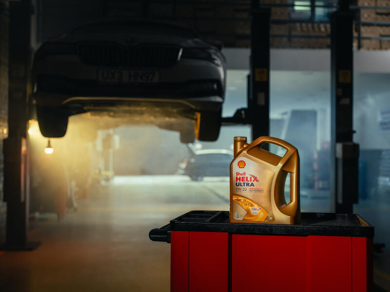 In a car repair shop, a car is lifted on a lift, and a container of Shell Helix Ultra 0W-20 motor oil is displayed on a red toolbox.