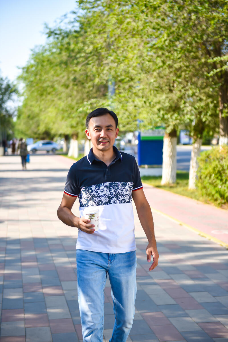 A man in a patterned polo shirt and jeans is walking along a paved path while holding a cup.