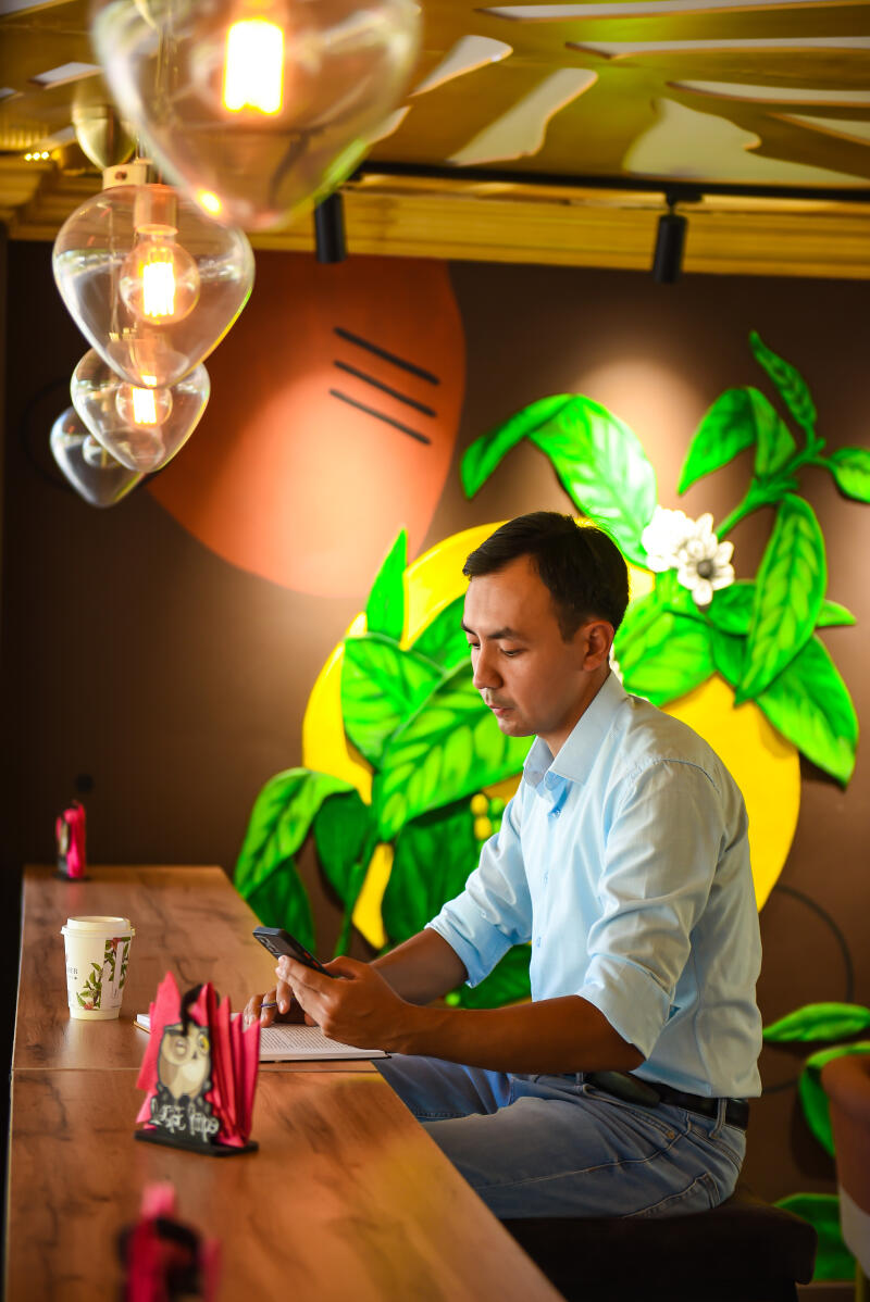 A man in a light blue shirt and jeans sits at a wooden table, looking at a smartphone in his hands, with a coffee cup and a napkin holder nearby; the background features a decorative wall with painted leaves and circles, and heart-shaped light bulbs hang from the ceiling.