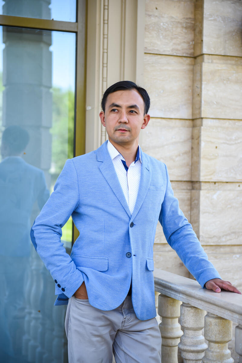 A man in a blue blazer and khaki pants stands with one hand in his pocket and the other resting on a stone railing.