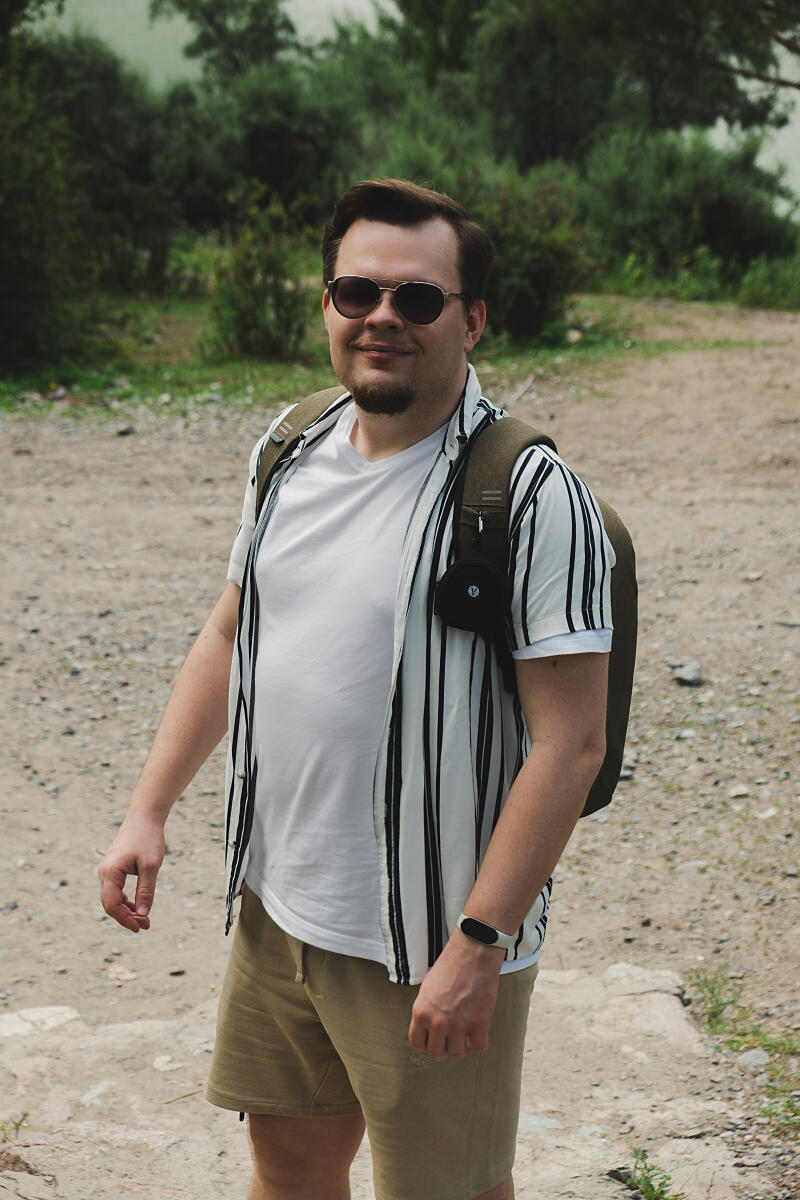 A man with sunglasses, a white t-shirt, a vertically striped shirt, and tan shorts stands with a backpack on a dirt path surrounded by greenery.