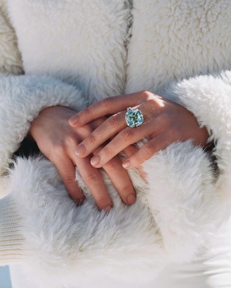 A person wearing a white furry coat has their hands clasped in front of them, with a large blue-green gemstone ring on their left ring finger.