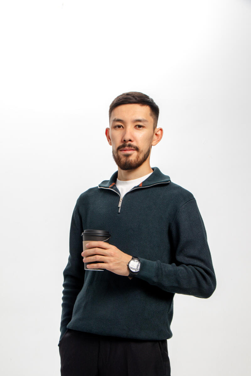 A man with a beard is holding a coffee cup, wearing a dark green sweater and a watch.