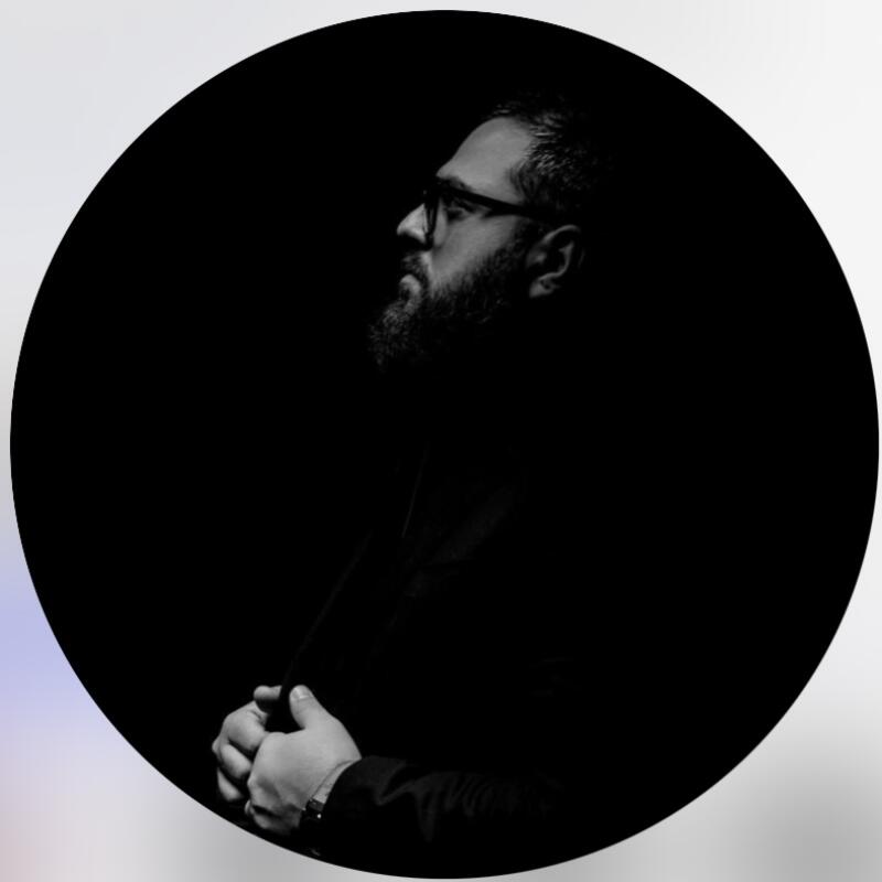 A black and white portrait of a man with glasses and a beard, looking off to the side, against a dark background.