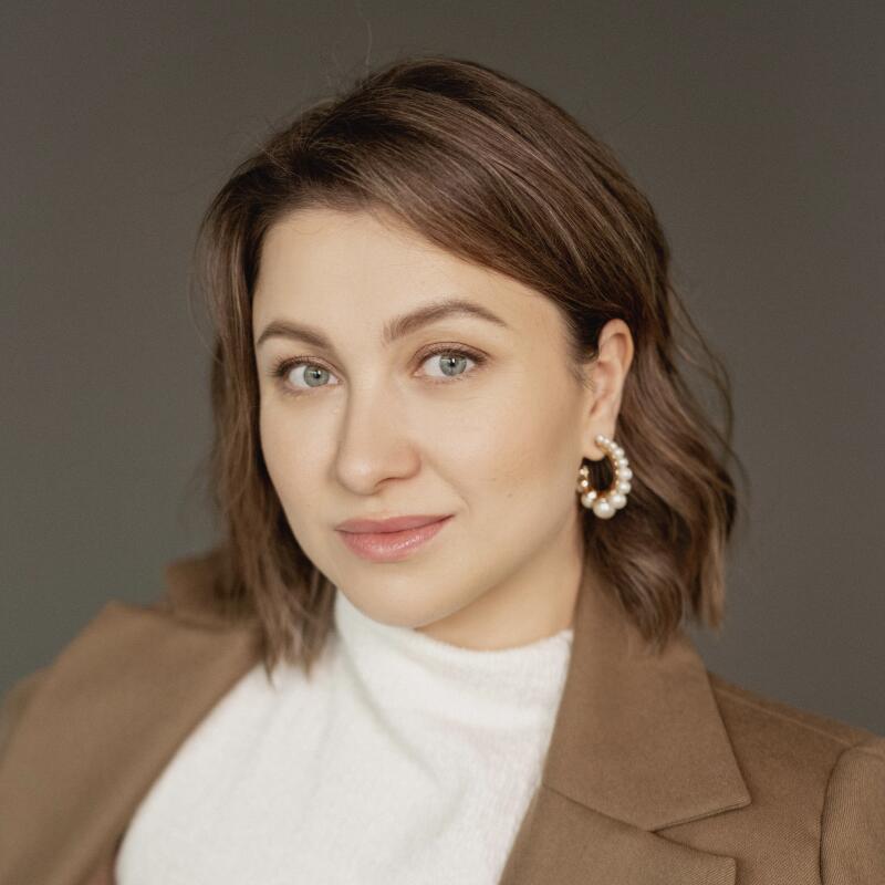 A woman with short brown hair, wearing a white turtleneck and a brown blazer, is posing with pearl earrings.