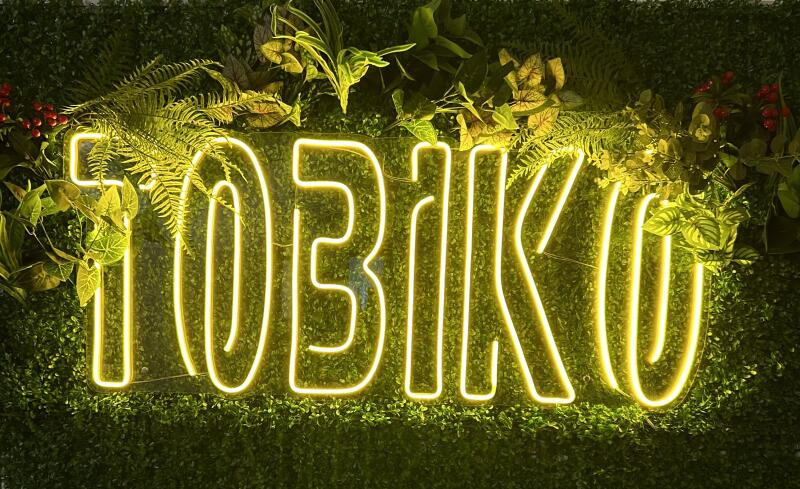 The image displays a neon sign spelling "TOBIKO" in bright yellow, set against a backdrop of dense green foliage.
