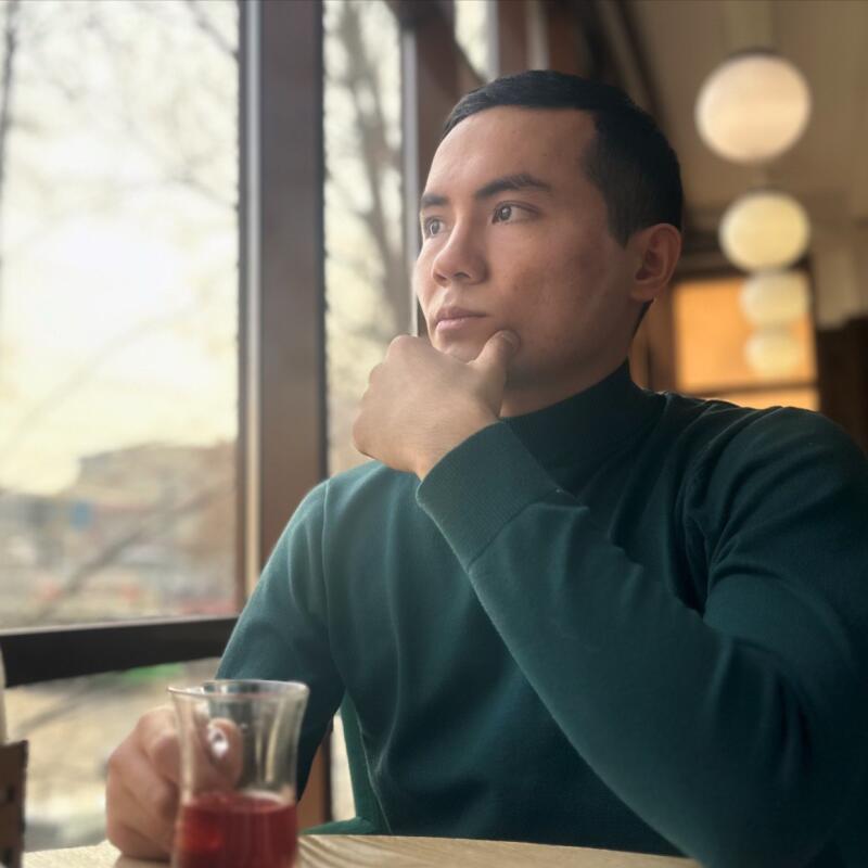 A man wearing a green turtleneck sweater is sitting at a table, looking out a window with his hand on his chin, with a glass of red liquid on the table.