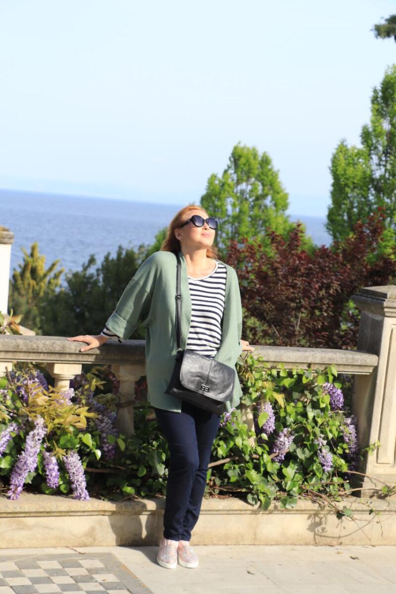 A woman with red hair, wearing sunglasses, a striped shirt, green cardigan, dark pants, and a black purse, is leaning against a stone railing with purple flowers.