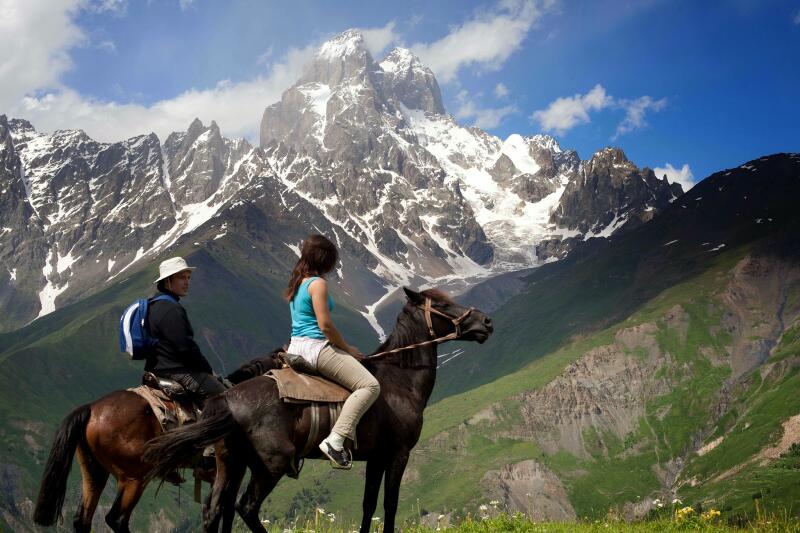 Two people are horseback riding in a mountainous area with snow-capped peaks and green hills.