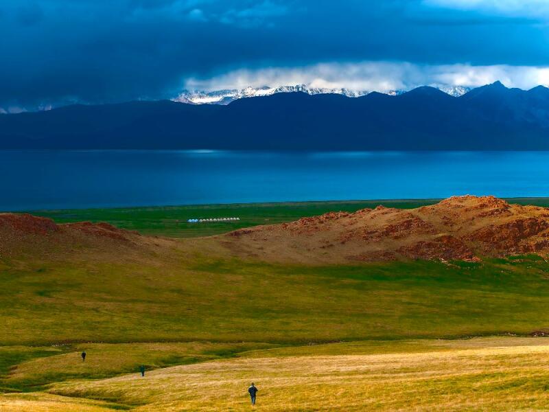 A scenic view of a field with people walking, a blue lake, and mountains in the background under a cloudy sky.