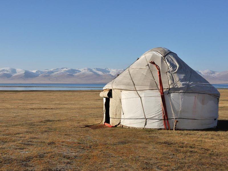 A white yurt stands in a field with mountains in the background under a blue sky.