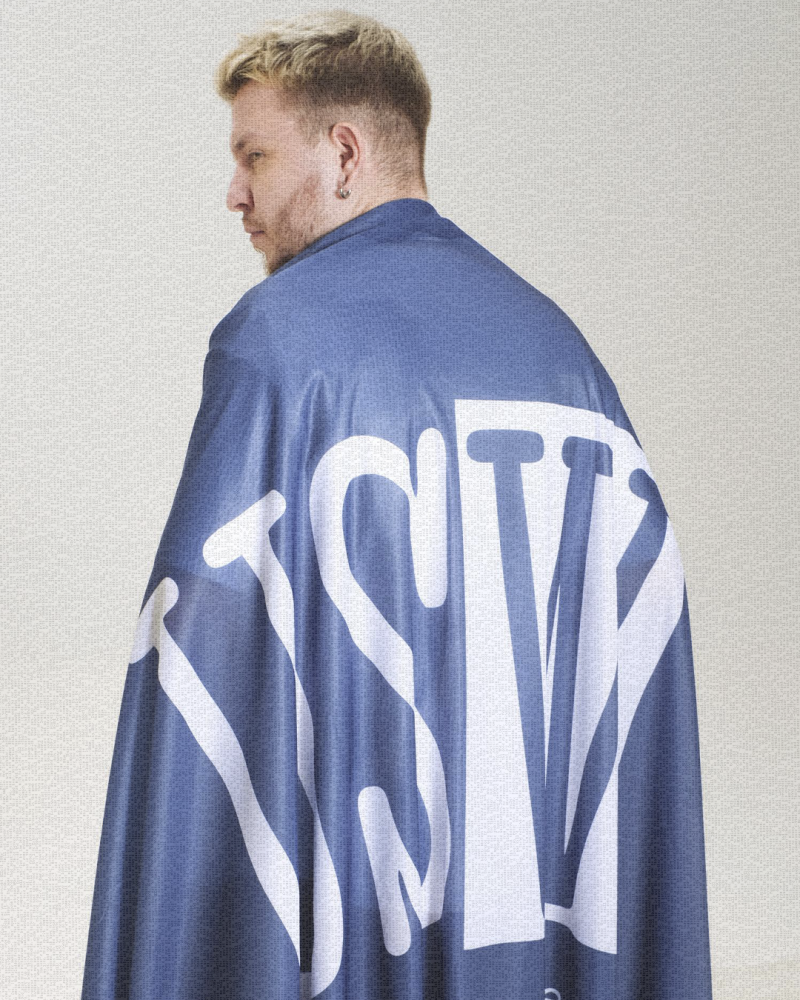 Мужчина со светлыми волосами стоит спиной к камере, на нем синий плащ с большими белыми буквами, складывающимися в слово «TSMV».