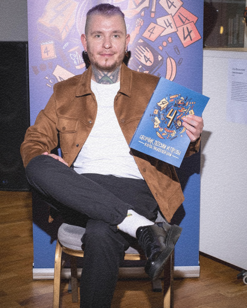 Татуированный мужчина с фиолетовыми волосами сидит на стуле и держит синюю книгу под названием «Сборник поэзии и прозы». На нем коричневая куртка, белая рубашка и черные брюки. На заднем плане синий баннер с цифрой 4 и различными иллюстрациями.