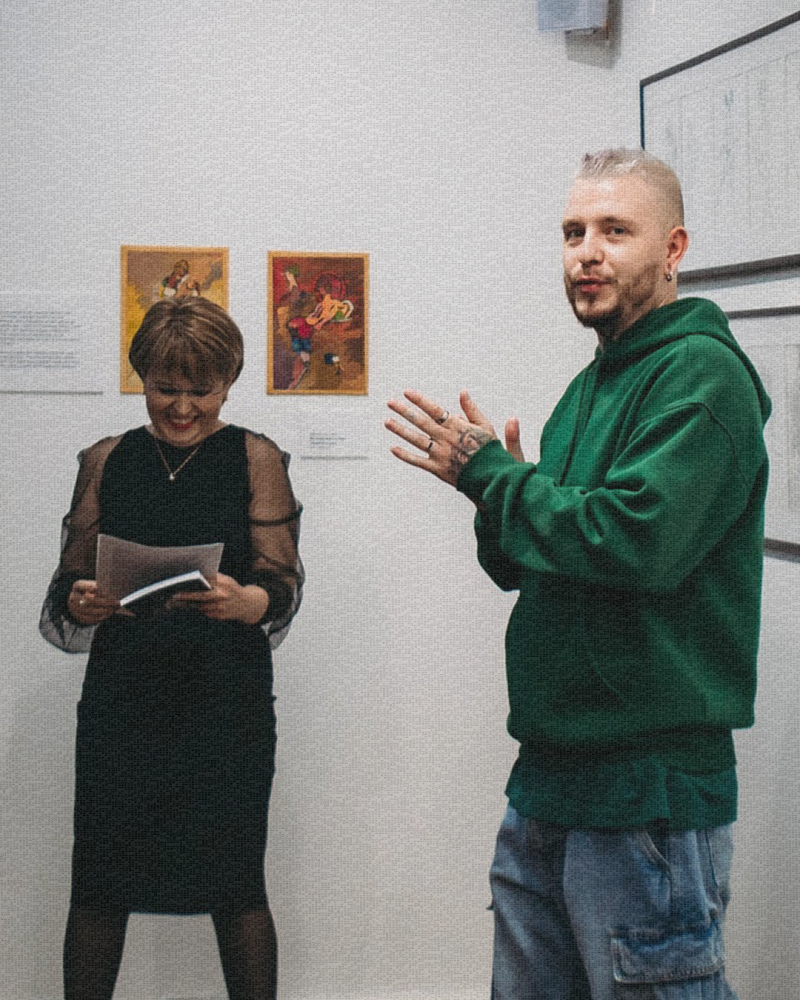 Женщина в черном платье держит блокнот и улыбается, в то время как мужчина в зеленой толстовке жестикулирует руками перед белой стеной, украшенной двумя картинами.