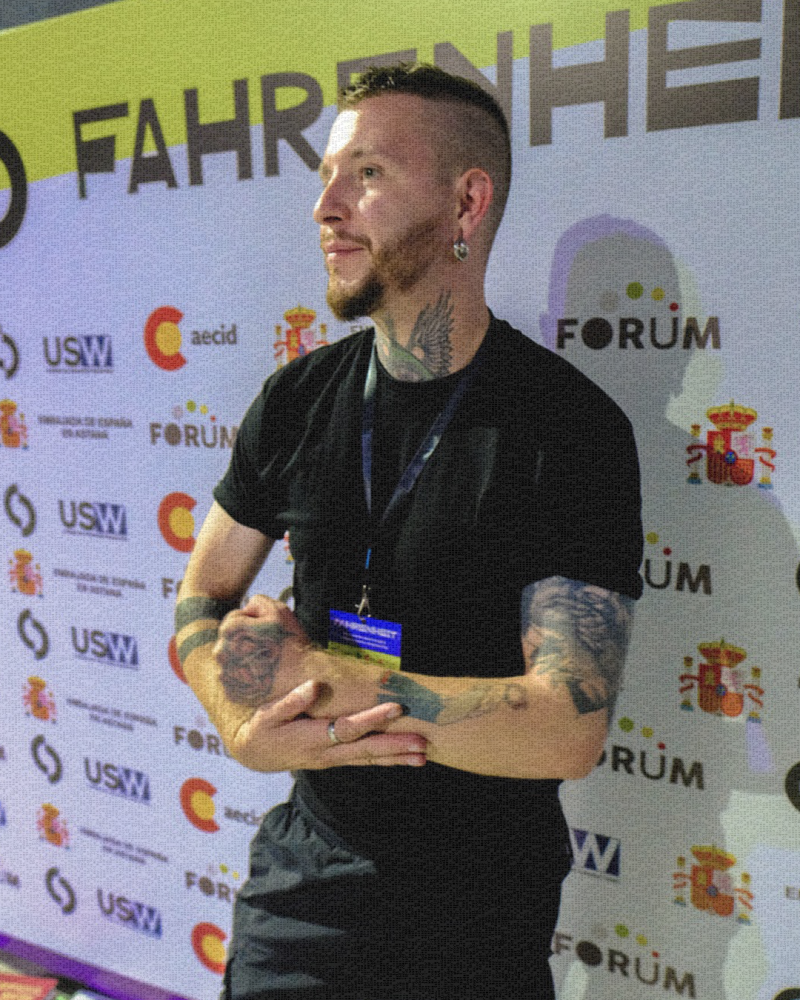 Мужчина с татуировками и в черной футболке стоит, скрестив руки, на фоне с логотипами и словом «FORUM».