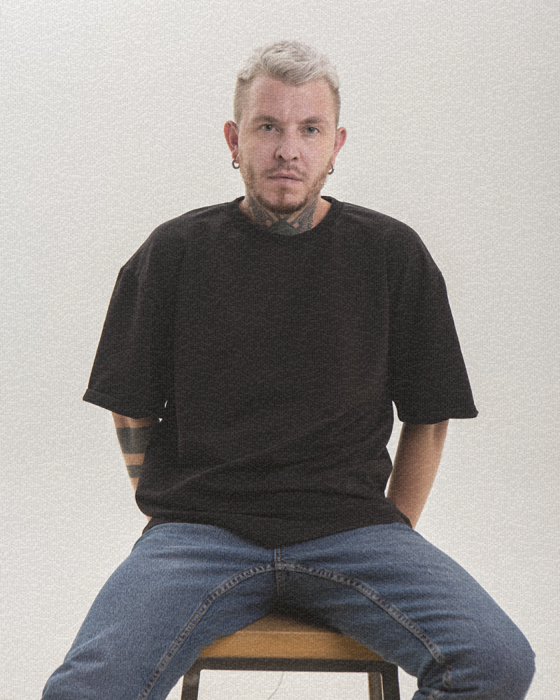 Человек с короткими белыми волосами и множеством татуировок сидит на деревянном стуле, одетый в черную футболку и синие джинсы.