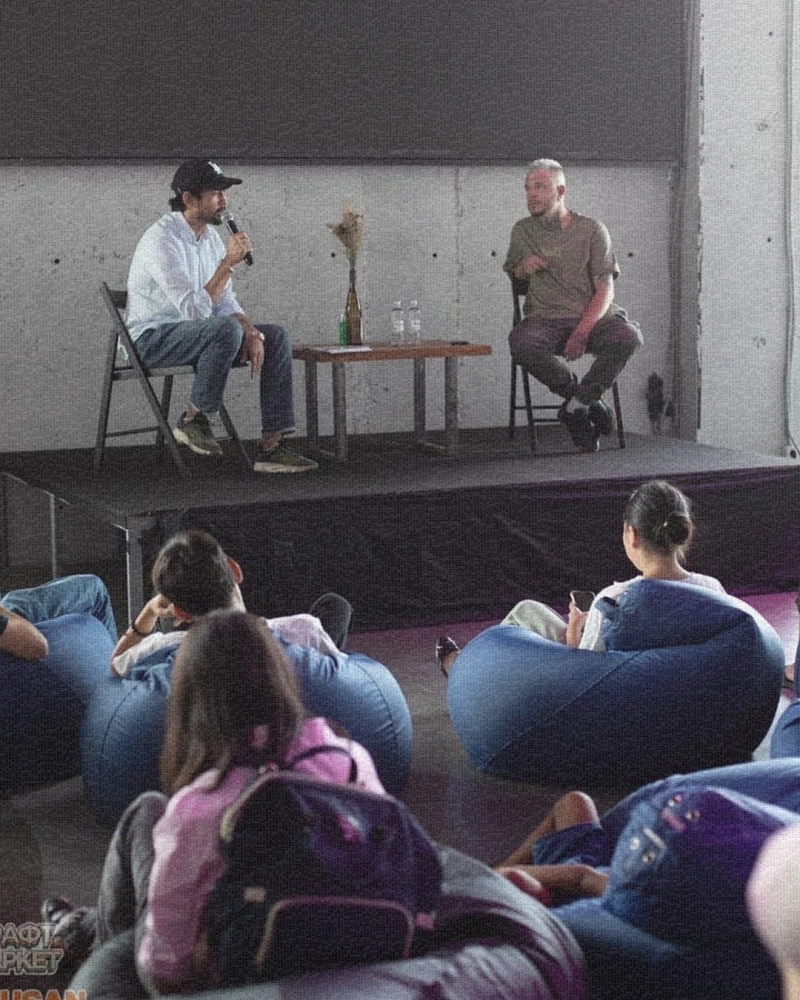 Два мужчины сидят на стульях на сцене, один держит микрофон, а зрители сидят на креслах-мешках и смотрят на них.