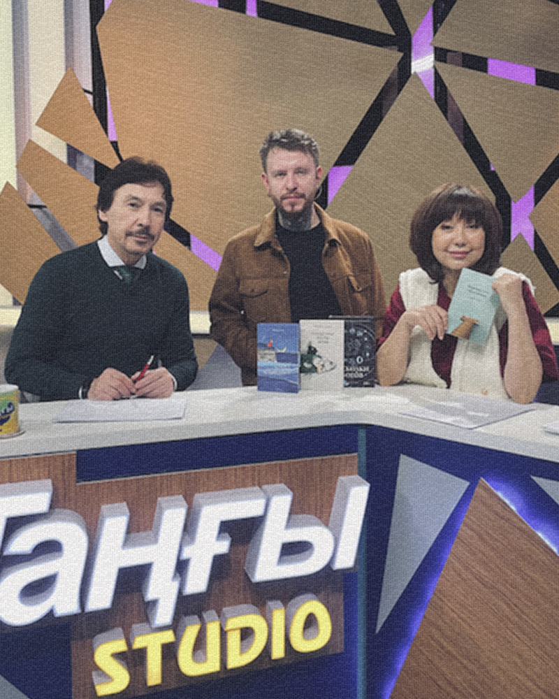 Три человека сидят за столом с книгами на фоне задника, на переднем плане виден текст "Ганғы STUDIO".