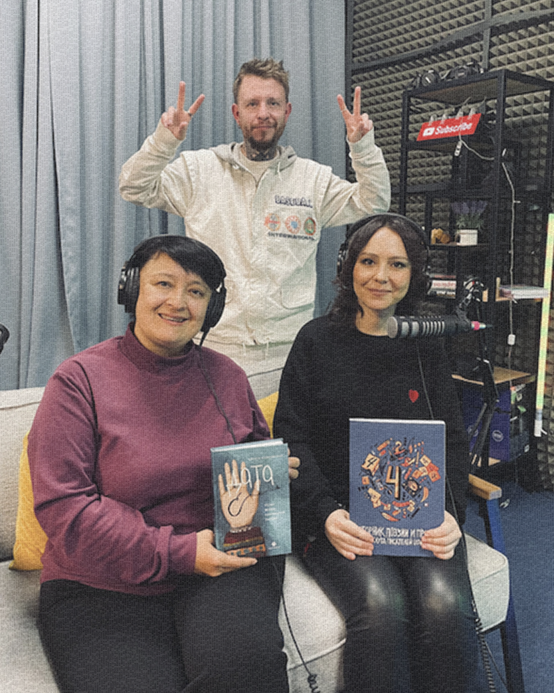 Три человека позируют в студии, держа в руках книги; мужчина на заднем плане показывает знак мира над ними. Женщина слева в наушниках и бордовой водолазке, а женщина справа в черном свитере с маленьким красным сердцем держит книгу под названием «4». На заднем плане видна надпись «Подписаться».