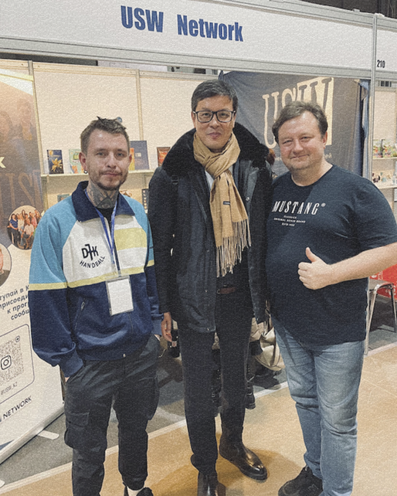 Три мужчины стоят перед стендом с надписью "USW Network"; мужчина слева одет в сине-белую куртку со словом "HANDBALL", мужчина посередине в очках и шарфе, а мужчина справа в черной футболке со словом "MUSTANG" и показывает большой палец вверх.